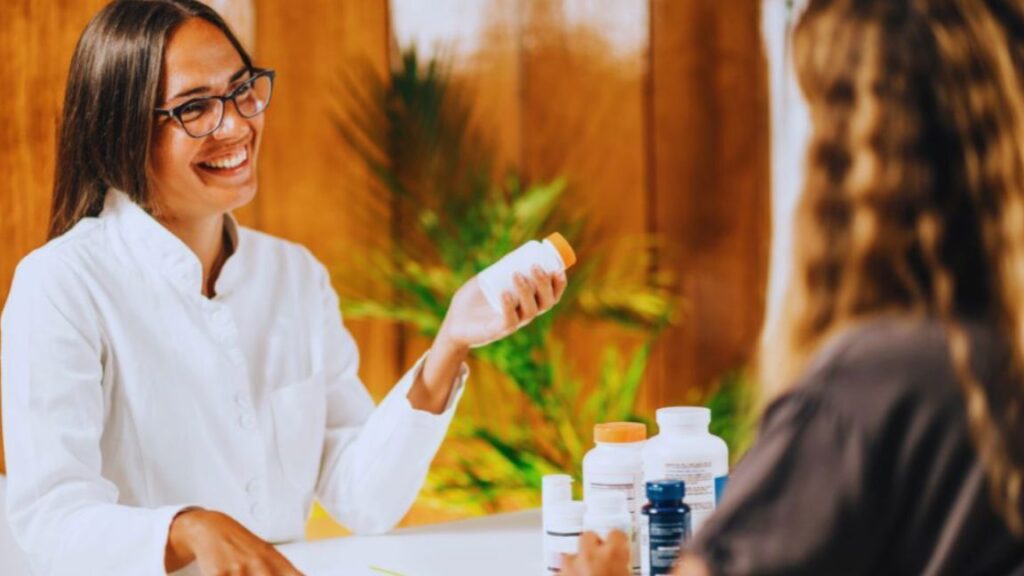 A functional medicine doctor in a white coat discusses supplements with a patient across a table, with several supplement bottles visible in a warmly lit office.