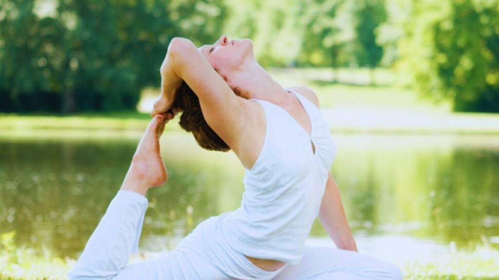 A person dressed in white performs an advanced yoga pose outdoors by a serene lake, surrounded by lush green trees, illustrating a peaceful moment of mindfulness and stress management.