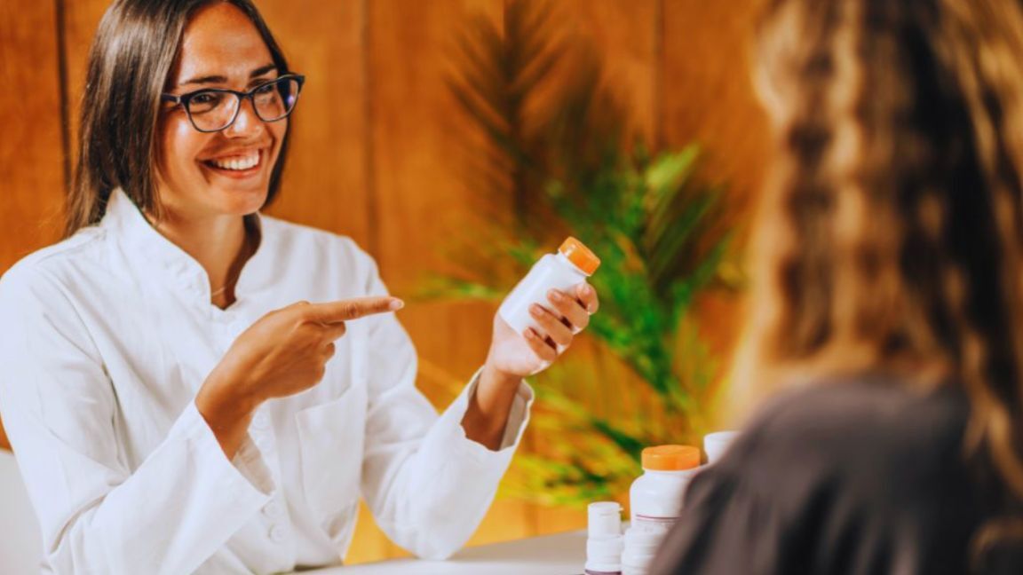 What Are Recovery Peptides And How Do They Work For Healing? 3 A doctor wearing a white coat discusses functional medicine near me options with a patient, pointing to a pill bottle in a warm, wood-paneled office with green plants in the background.