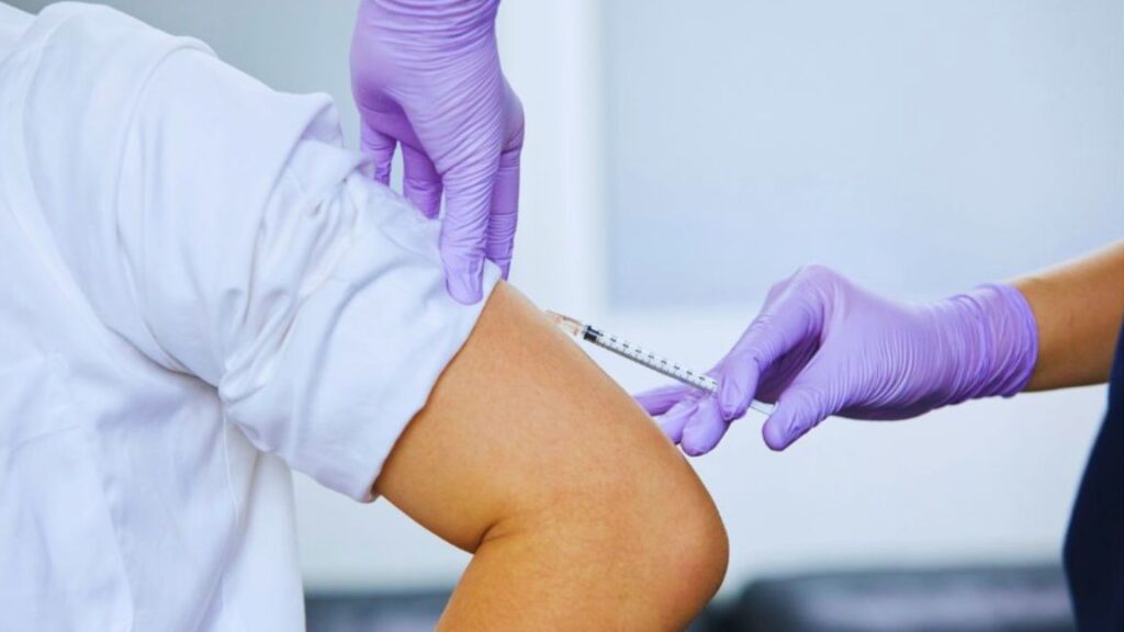 A person in a white shirt receives an injection in the upper arm from someone wearing purple medical gloves. The scene suggests a clinical setting where treatment or supplementation—potentially involving muscle recovery peptides—is being administered.