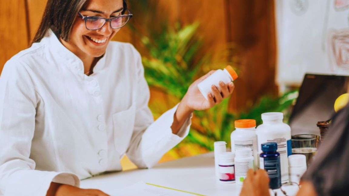 A functional medicine doctor wearing a white coat smiles while discussing supplement bottles with a patient, holding one bottle in her hand at a desk filled with various vitamins and containers.