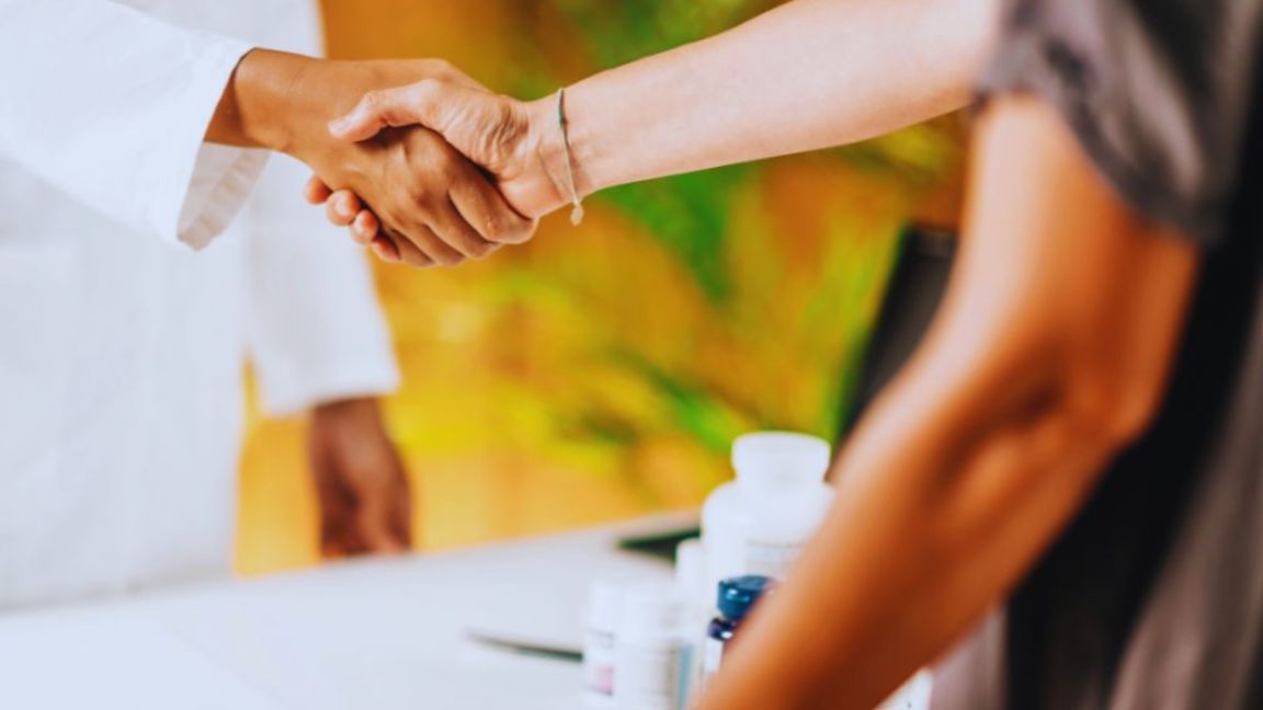 A functional medicine doctor in a white coat shakes hands with a patient at the end of a consultation, with supplement bottles visible on the table in the foreground.