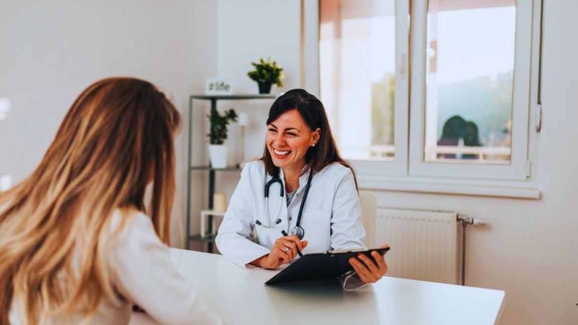 A female functional medicine doctor in a white coat sits at a desk holding a digital tablet and speaking with a female patient across from her in a bright medical office with large windows and shelves in the background.