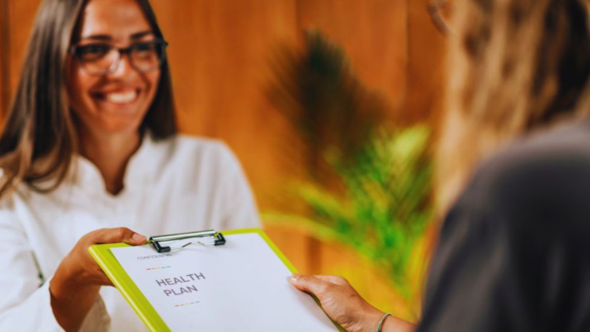 A functional medicine Aventura professional hands a clipboard labeled “Health Plan” to a patient in a warm, wood-paneled office with a green plant in the background.
