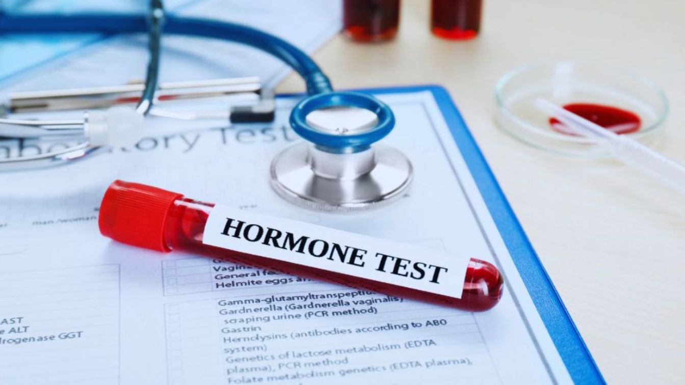 Blood sample tube labeled “hormone test” lying on a laboratory test form with a stethoscope and other blood samples in the background, representing hormone testing for possible hormone imbalance.