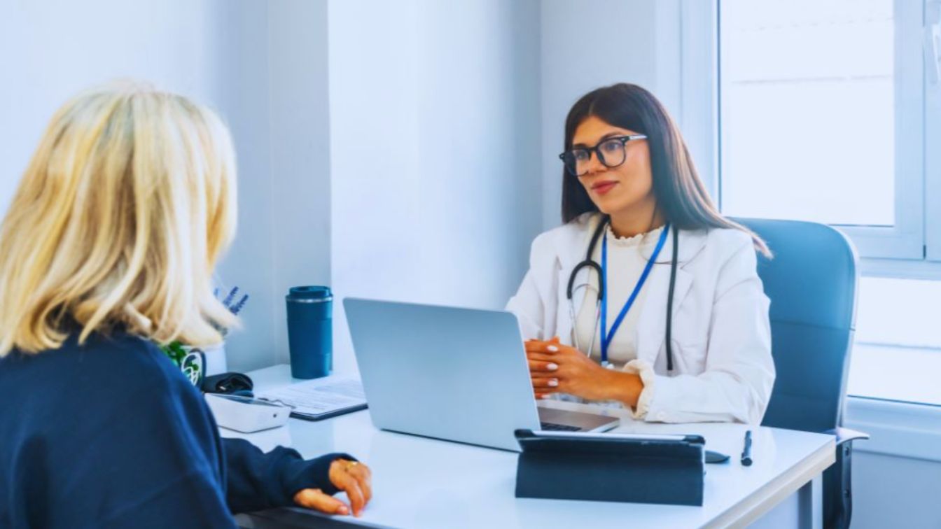 Holistic doctor in a white coat sitting at a desk with a laptop and stethoscope, talking with a middle-aged female patient in a bright Miami medical office.