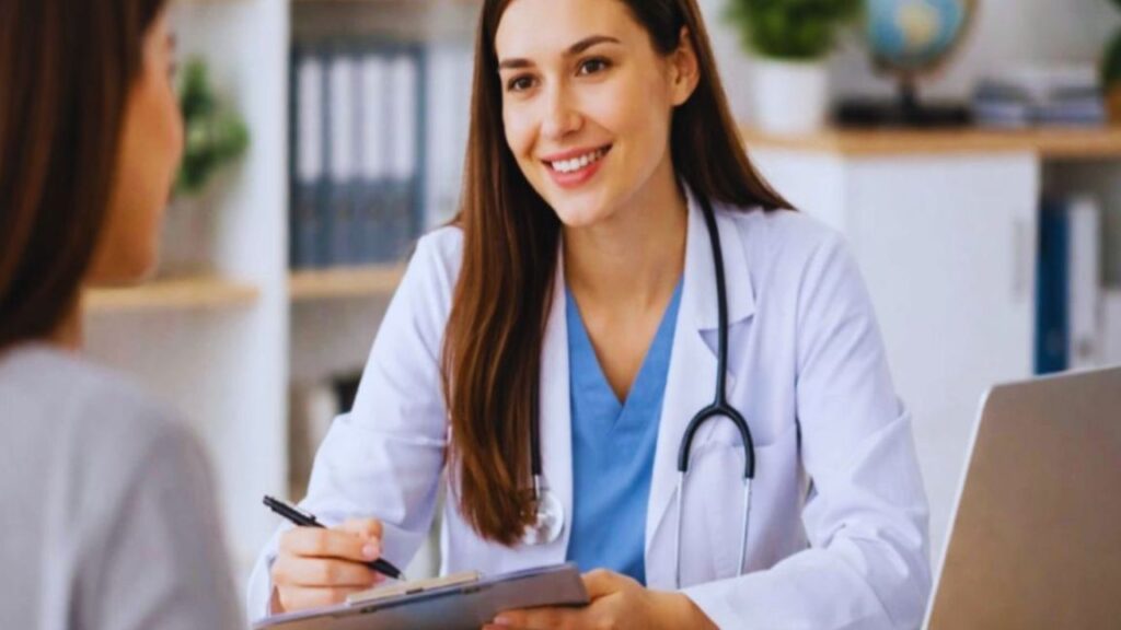 female internal medicine doctor in a white coat sits at a desk in a modern medical office, holding a clipboard and pen while attentively speaking with a seated patient in the foreground, symbolizing personalized internal medicine care in Aventura, FL.