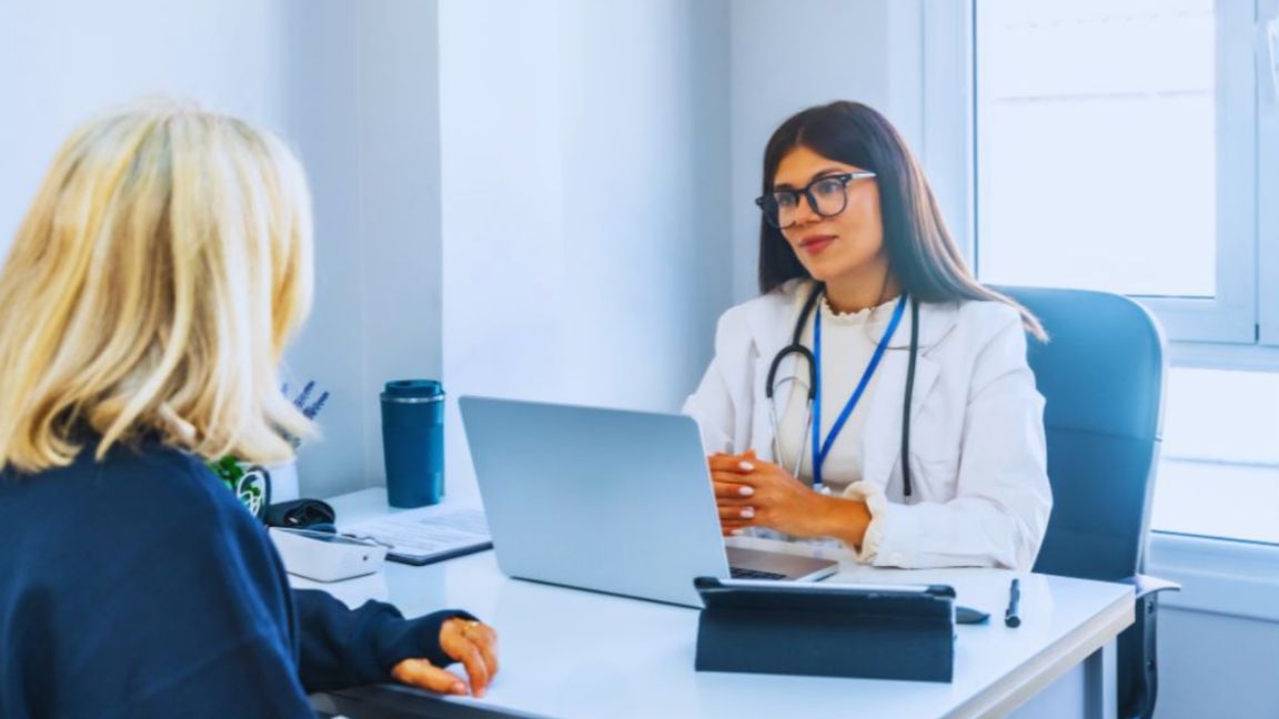 Internal medicine doctor with stethoscope in a white coat consults with a blonde female patient at a desk in a contemporary office, using a laptop and tablet during a checkup in Aventura, FL.