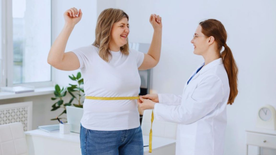 A woman celebrates as a healthcare provider measures her waist with a tape measure during a weight loss consultation in a bright medical office.