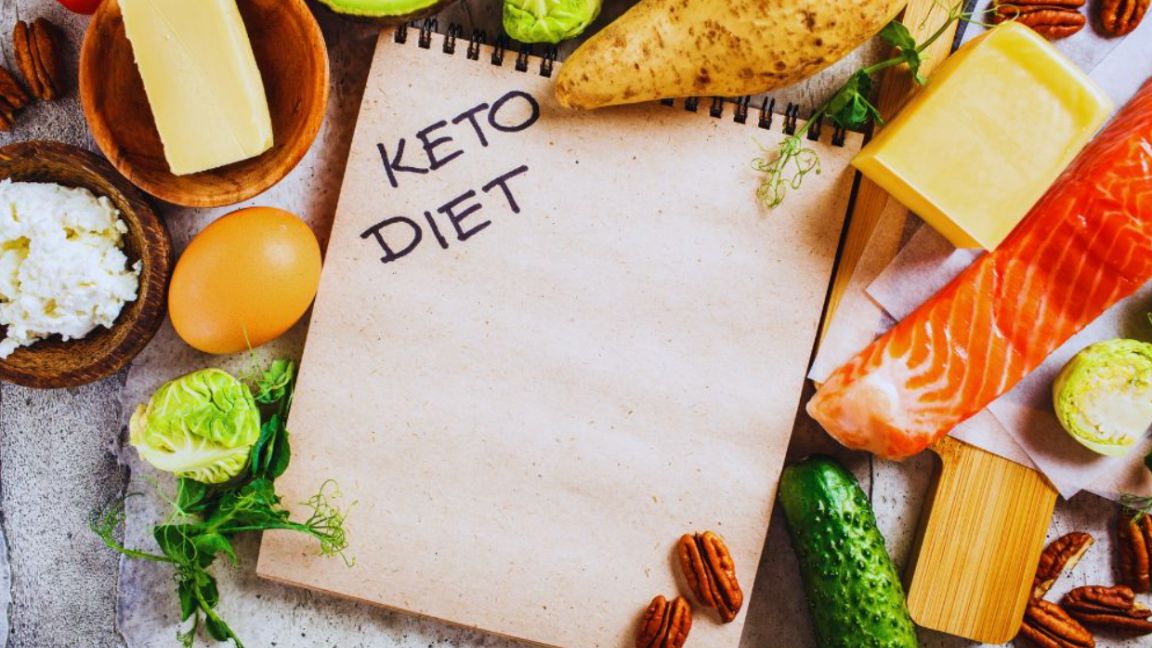 A view of a blank notepad labeled “Keto Diet” surrounded by low-carb foods including salmon, cheese, butter, eggs, nuts, cucumber, leafy greens, and avocado.
