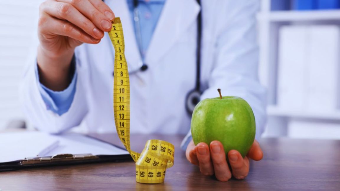 Close-up of a doctor in a white coat holding a tape measure in one hand and a green apple in the other, symbolizing medical obesity and weight loss programs in Aventura, FL.