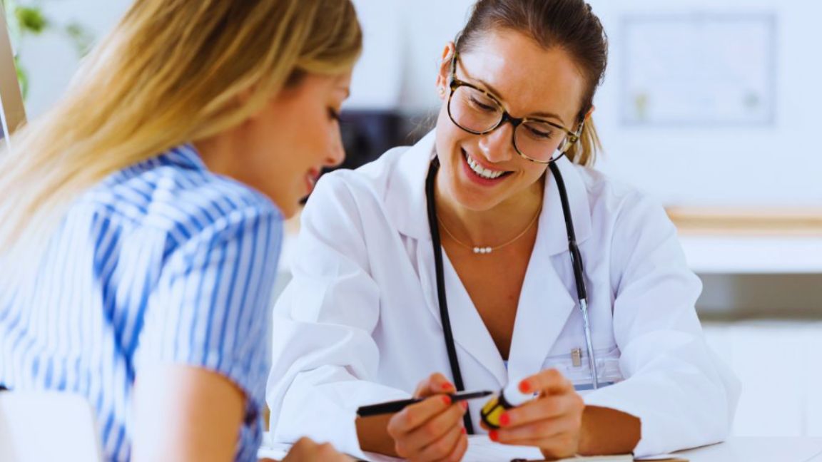 A female primary care doctor is discussing health concerns with a patient in a clinical setting.