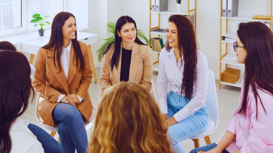 Adult women in a wellness support group seated in a bright office, engaged in supportive conversation.