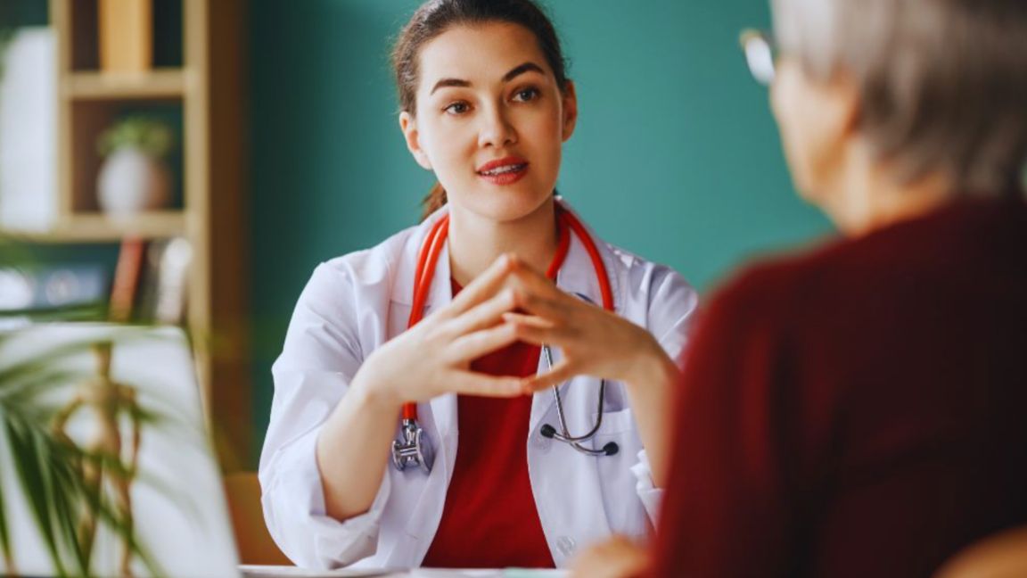 Physician in white coat with red stethoscope discussing an integrative health care plan with an older patient in a calm, modern clinic setting, both faces blurred for privacy.